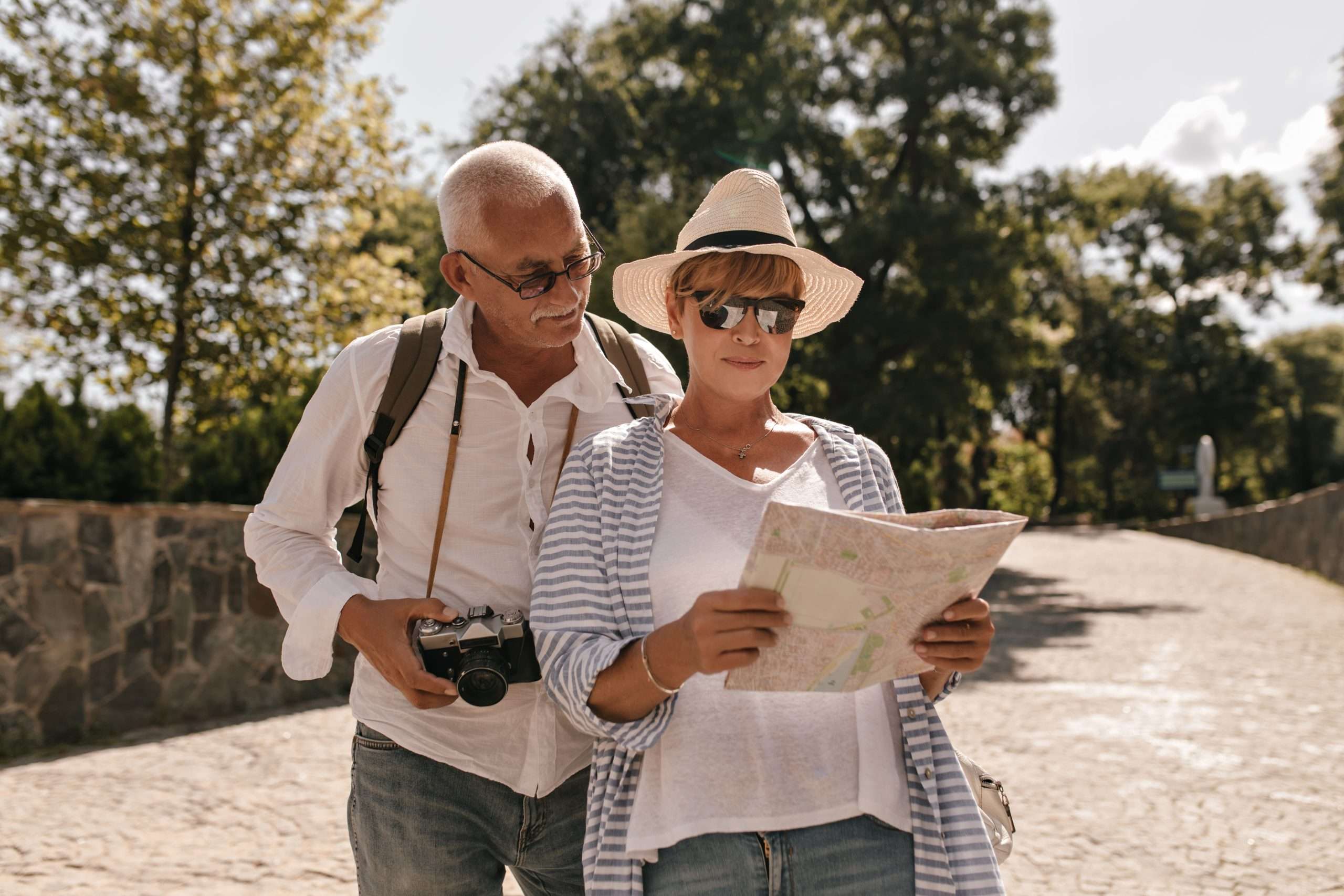 modern woman white tshirt striped blouse hat glasses looking map walking with grey haired man shirt with camera outdoor scaled